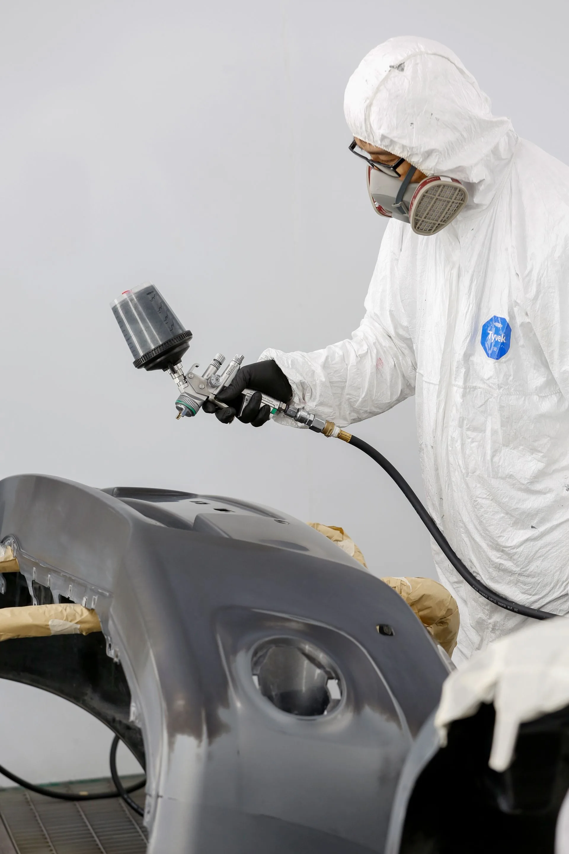 Technician in protective equipment refinishing a vehicle panel with a spray gun
